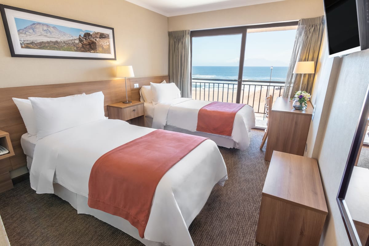 Een hotelkamer met twee bedden, een bureau, een televisie, een raam met uitzicht op de oceaan en een ingelijst schilderij aan de muur.
