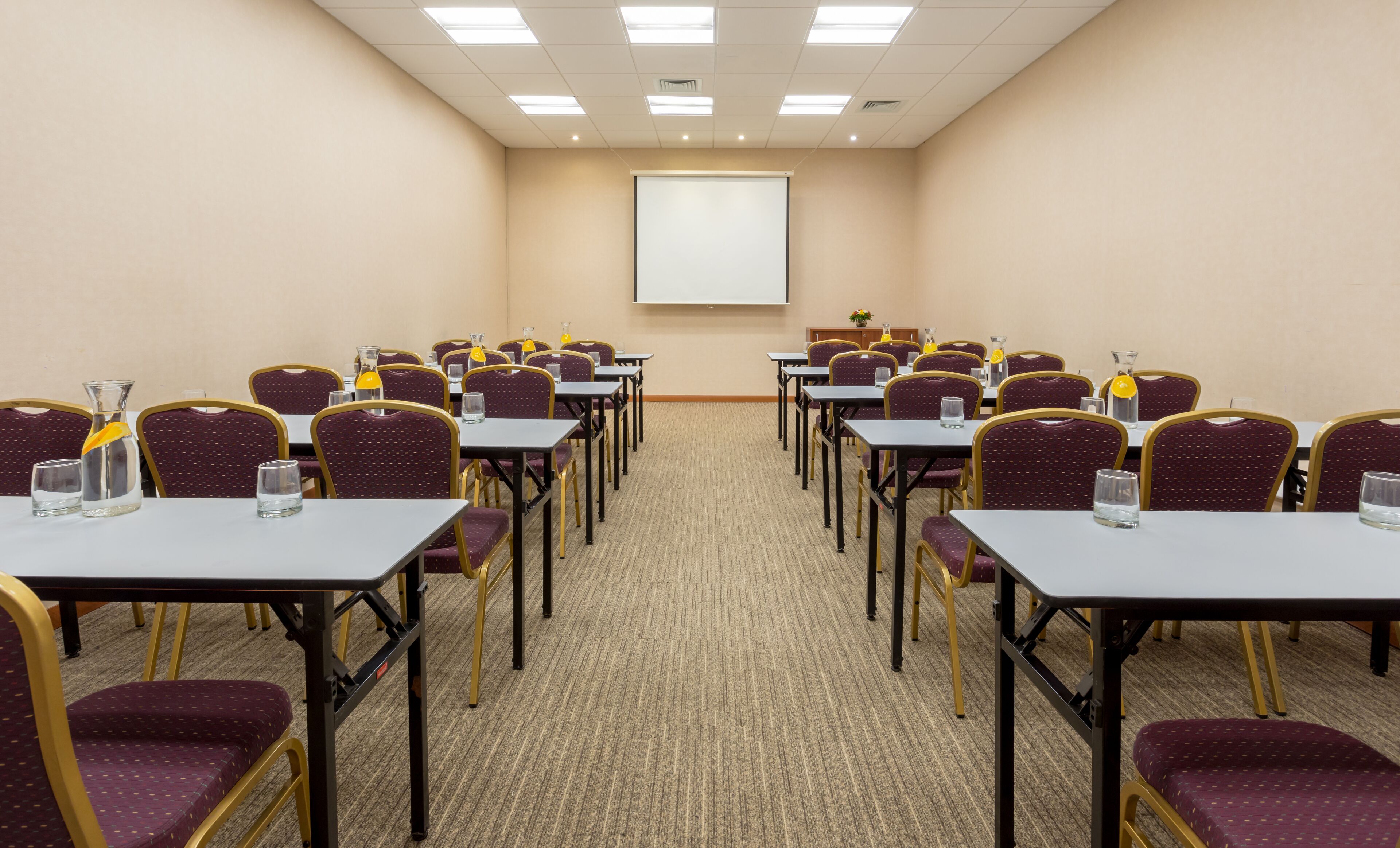 A conference room with rows of tables and chairs, a projector screen, and a carpeted floor.