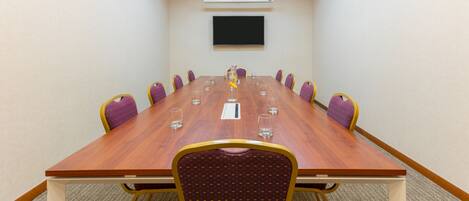 A conference room with a long wooden table, purple chairs, and a flat-screen TV on the wall.