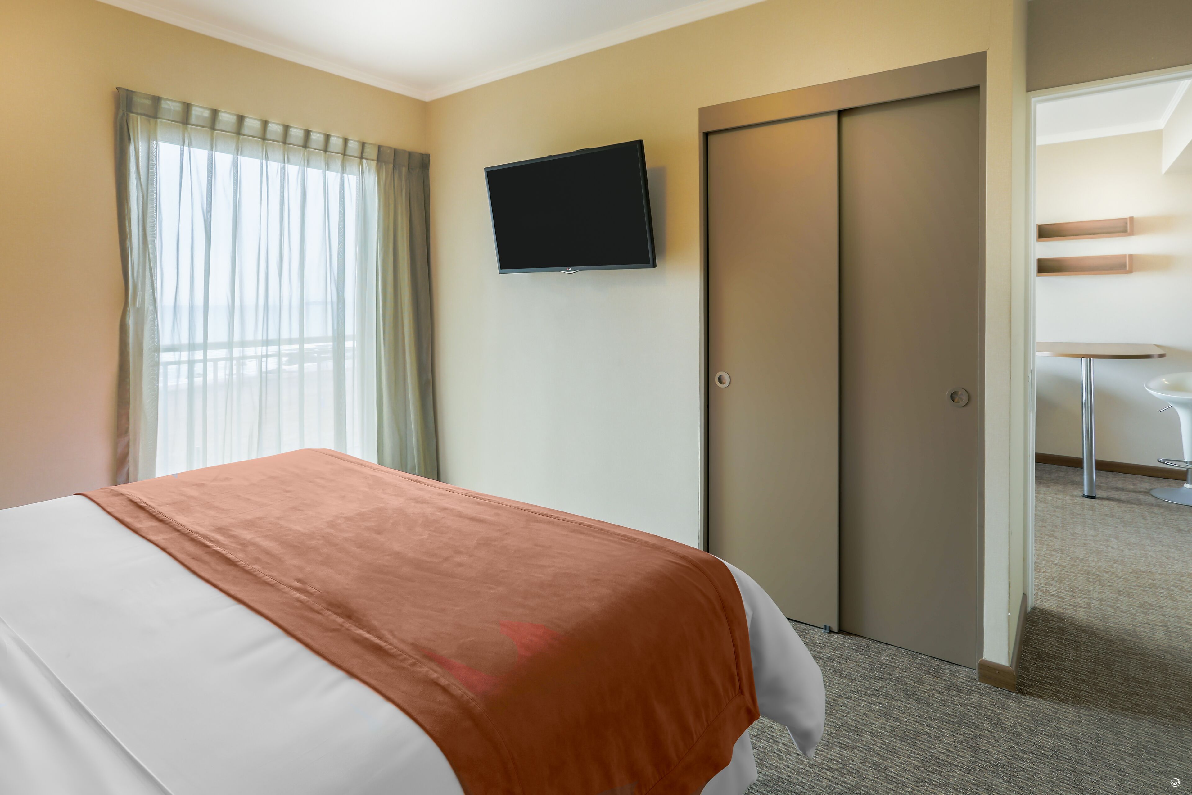 A hotel room with a bed, a television on the wall, a door, and a small table with a chair.