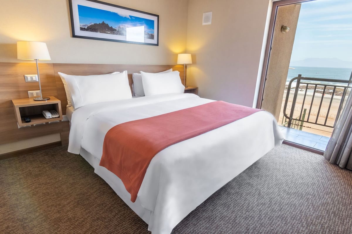 Een hotelkamer met een bed, nachtlampjes, een nachtkastje, een lijst aan de muur en uitzicht op zee door een balkondeur.