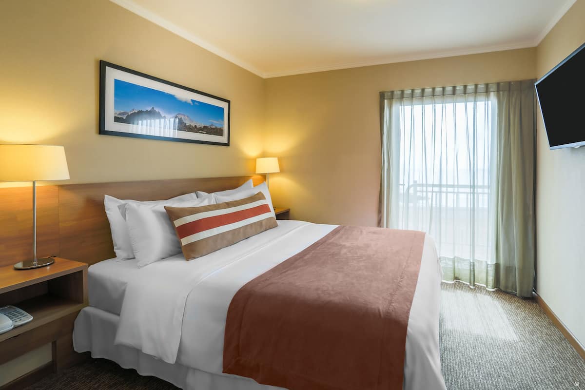 Hotelkamer met een bed, twee bedlampen, een wandtelevisie, een lijst aan de muur en een raam met gordijnen.