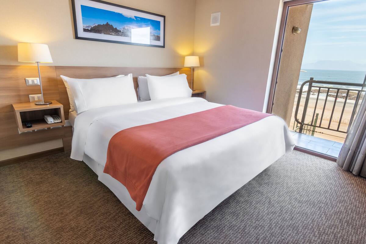 Hotelkamer met een bed, twee bedlampen, een nachtkastje met telefoon, een ingelijst schilderij aan de muur, en uitzicht op het strand door een geopende deur.