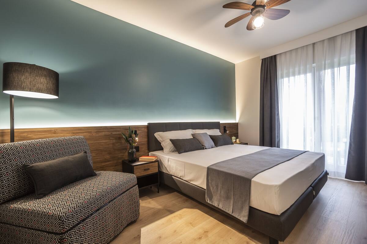 A modern hotel room with a large bed, a patterned armchair, a bedside table with a lamp, and a ceiling fan.