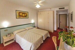 Habitación de hotel con dos camas, una mesita de noche, un armario, un ventilador de techo, un cuadro y una planta en maceta.