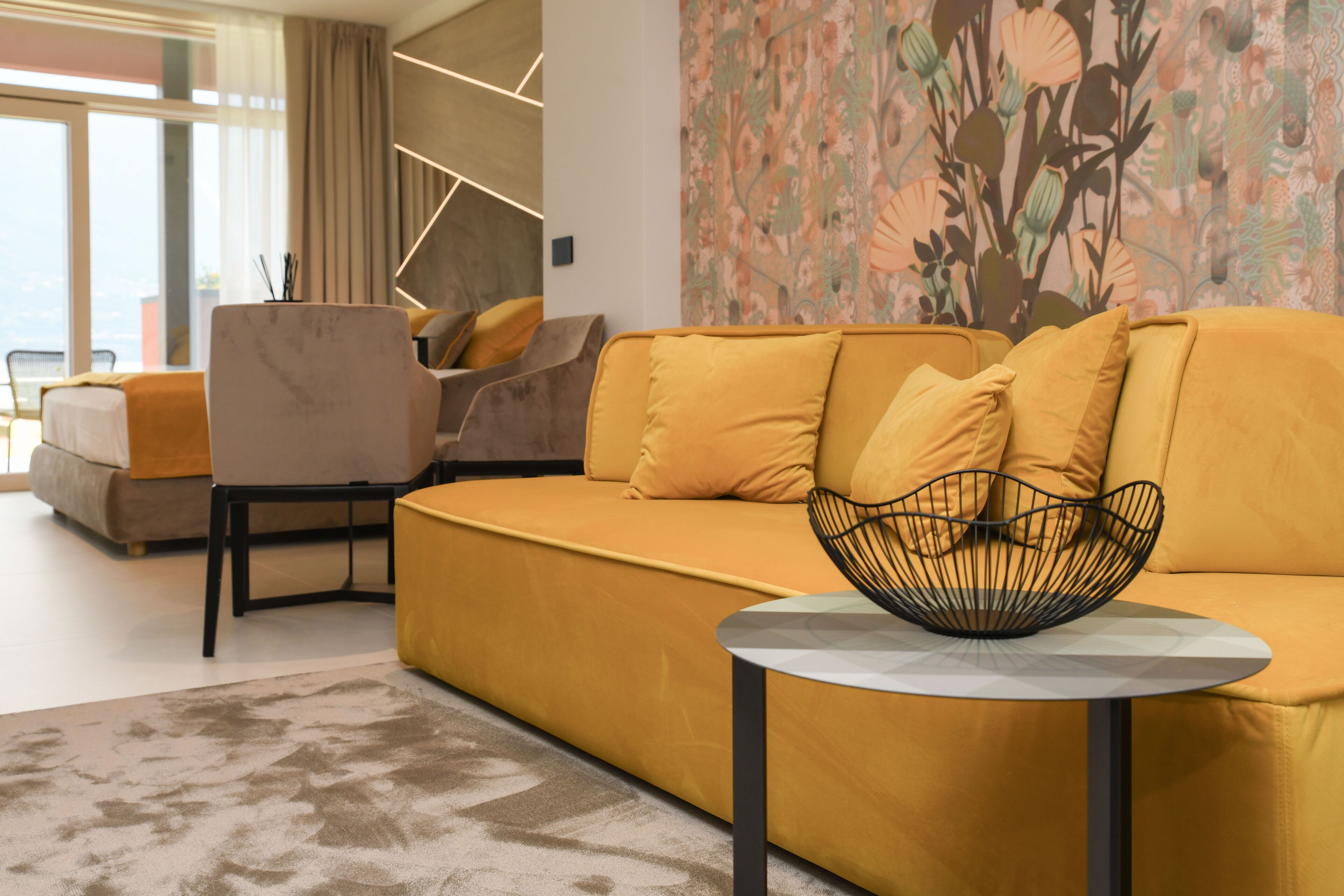 Ein modernes Wohnzimmer mit einem gelben Sofa, einem runden Couchtisch mit Drahtkorb und einer Wand mit Muster.