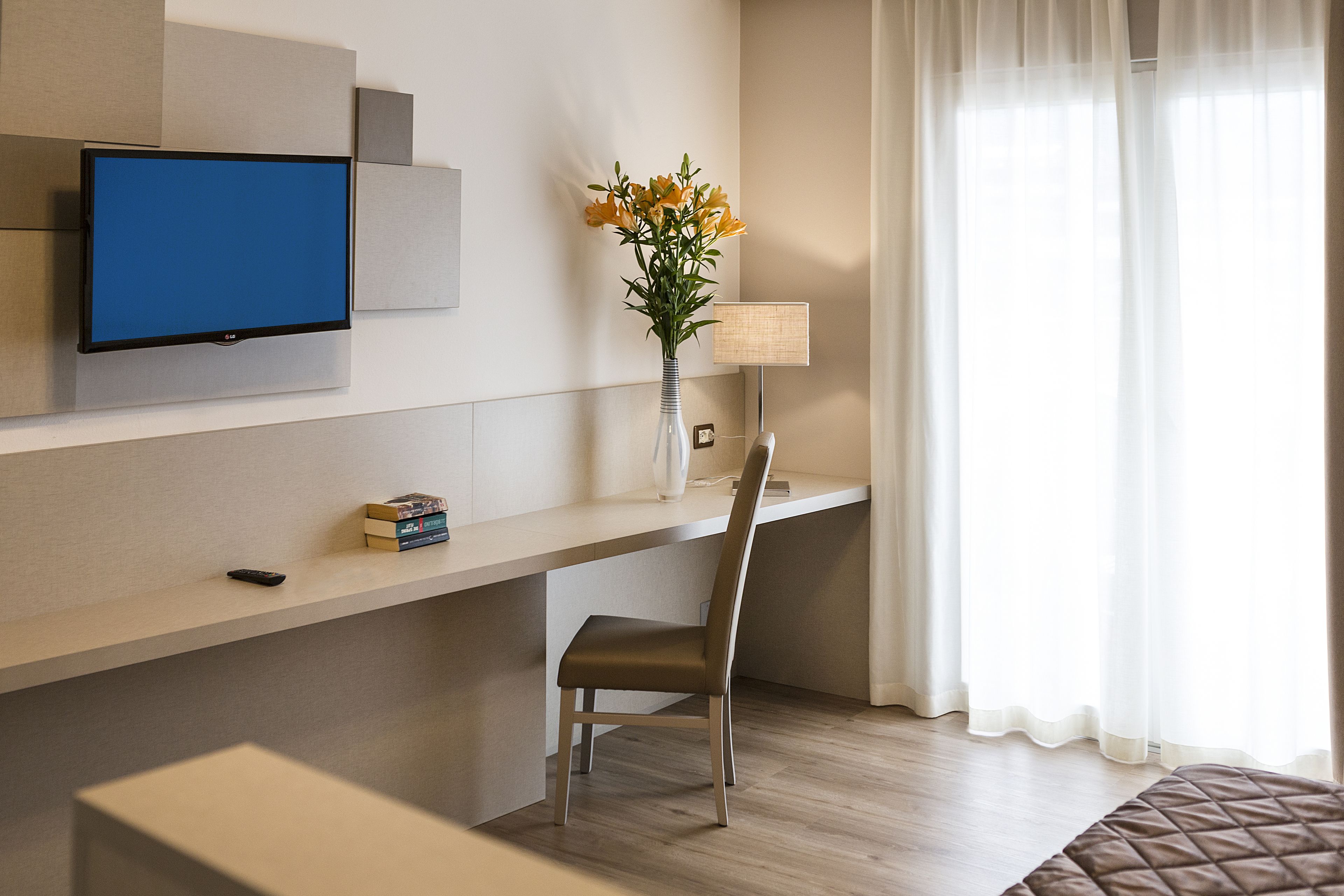 A modern hotel room with a flat-screen TV mounted on the wall, a desk with a chair, a vase with flowers, a lamp, and a large window with sheer curtains.