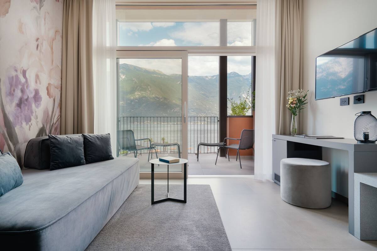 A modern living room with a sofa, a small round table, a desk with a chair, a television, and a view of mountains through large windows.