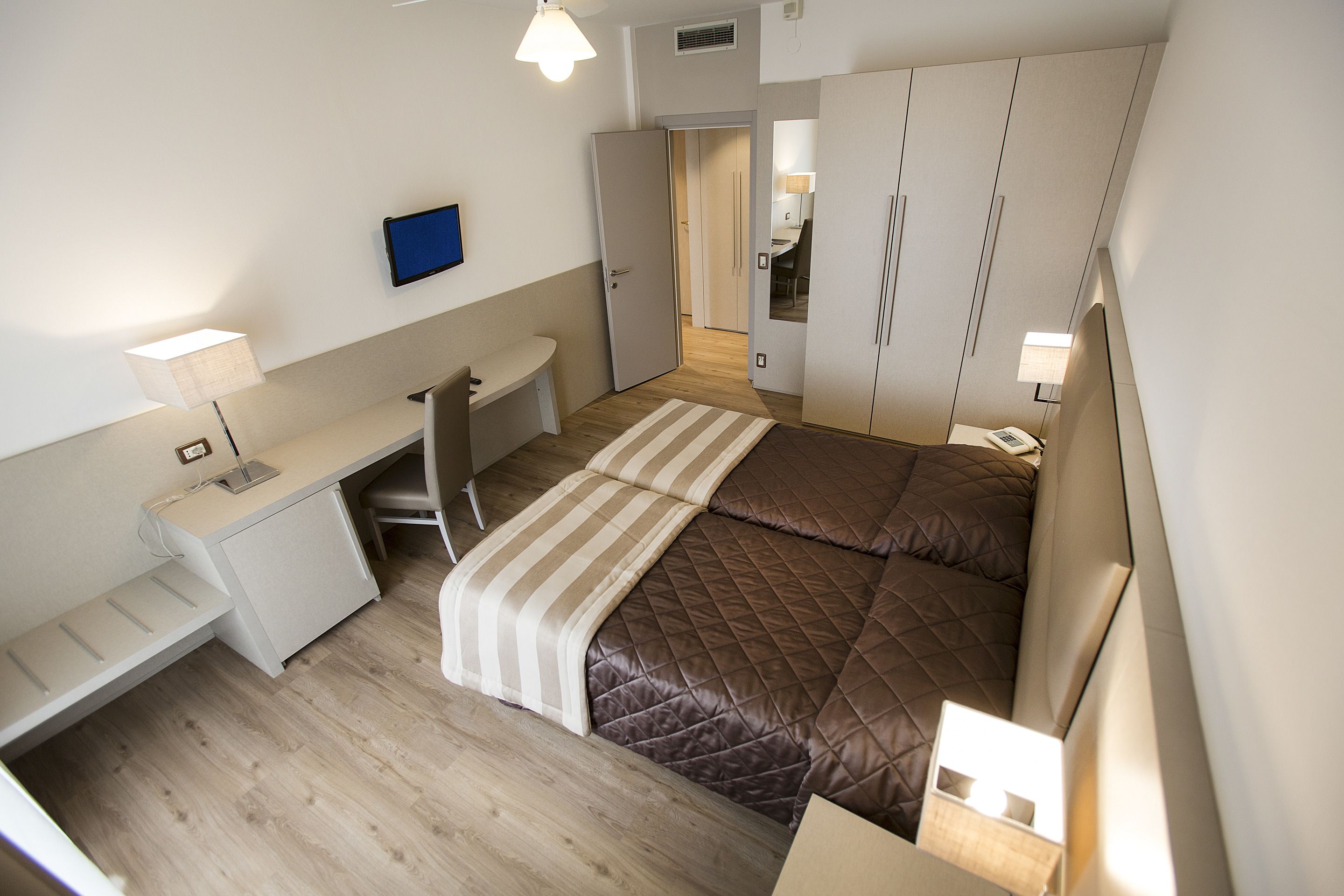 Un dormitorio compacto con una cama, un escritorio y una silla. Hay un televisor montado en la pared y un armario con espejo.