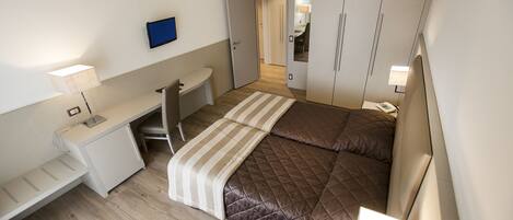 Un dormitorio compacto con una cama, un escritorio y una silla. Hay un televisor montado en la pared y un armario con espejo.