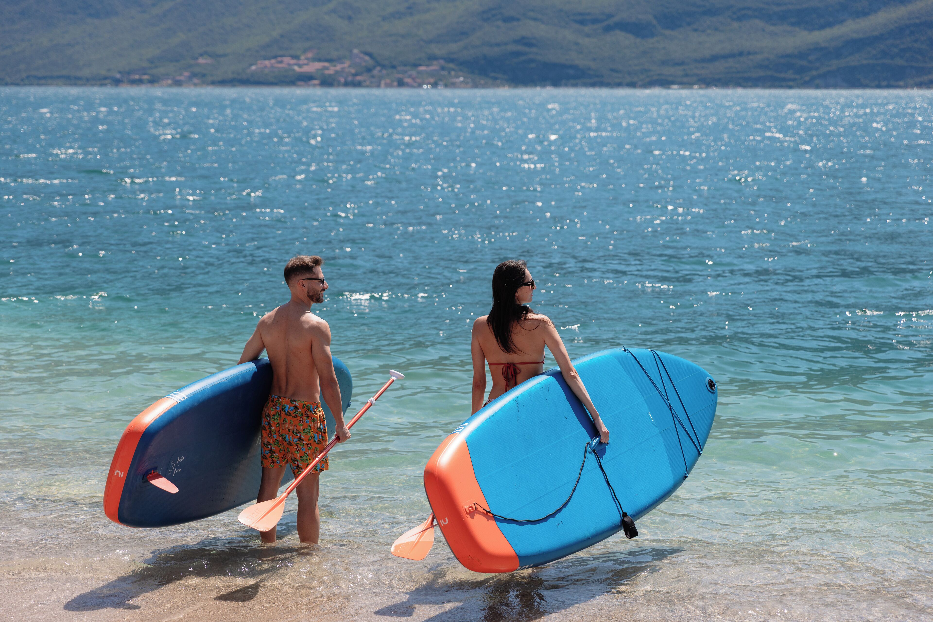 Deux personnes transportant des planches de paddle près d’une plage, avec une eau claire et des collines qui se dessinent à l’horizon.
