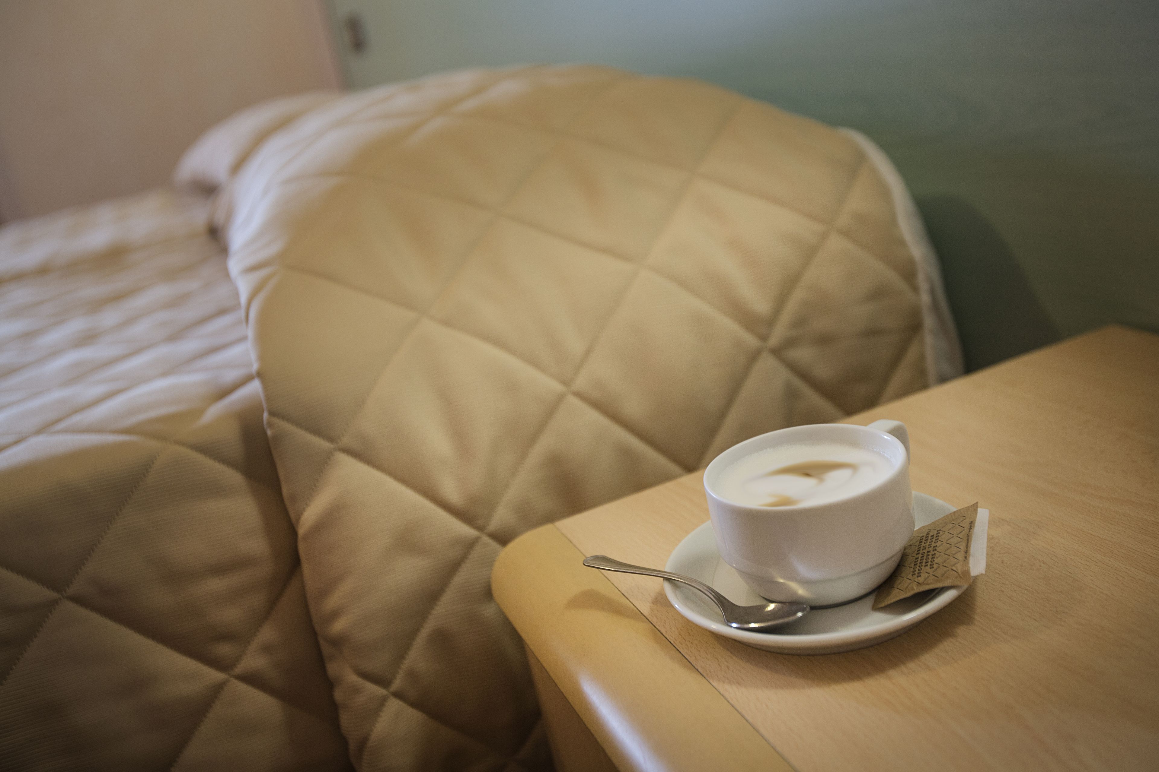 A cup of coffee on a table next to a bed with a quilted headboard.