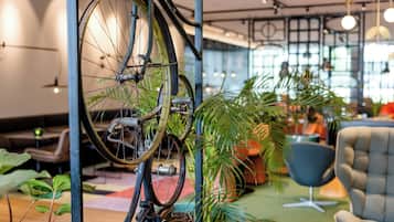 Ein Fahrrad, das an einer Metallstange in einem hellen Innenraum lehnt, mit großen Fenstern, Topfpflanzen und moderner Möbel.
