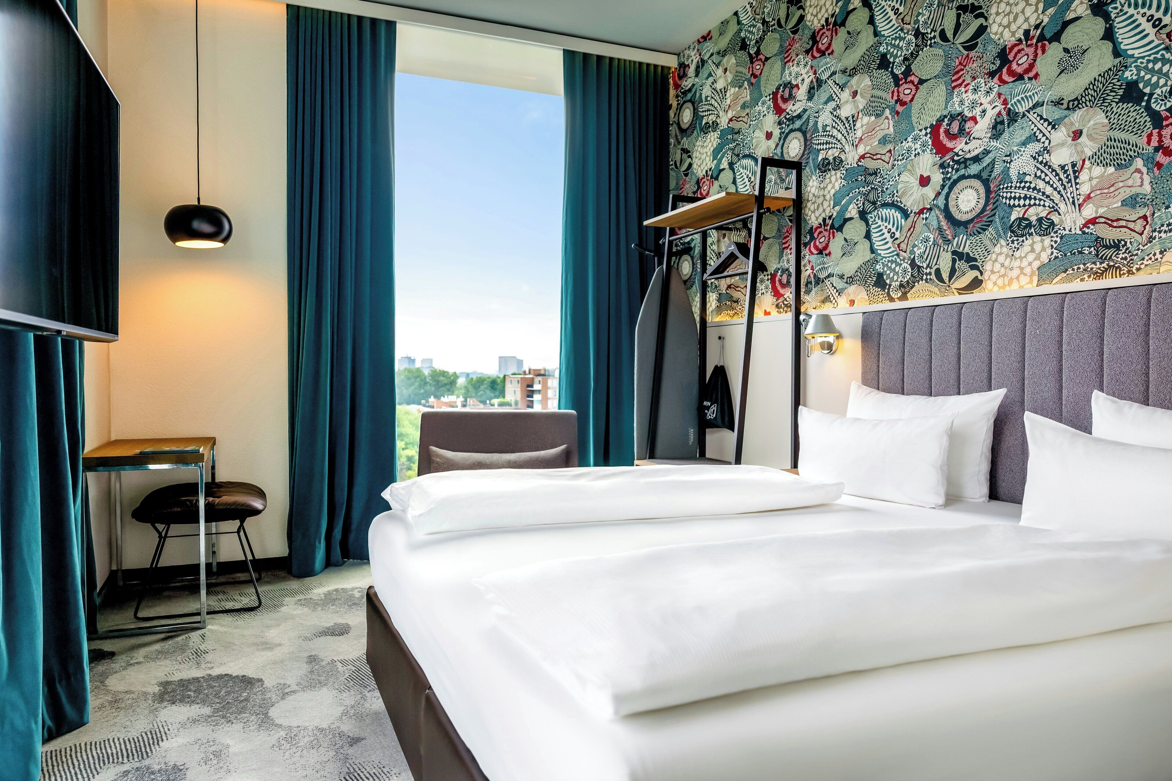 A hotel room with a large bed, a desk, a chair, a wardrobe, and a view of the city.