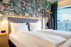 A hotel room with a large bed, a headboard with a patterned design, a wall-mounted lamp, and a window with a view of a building. - Motel One Amsterdam (Amsterdam)