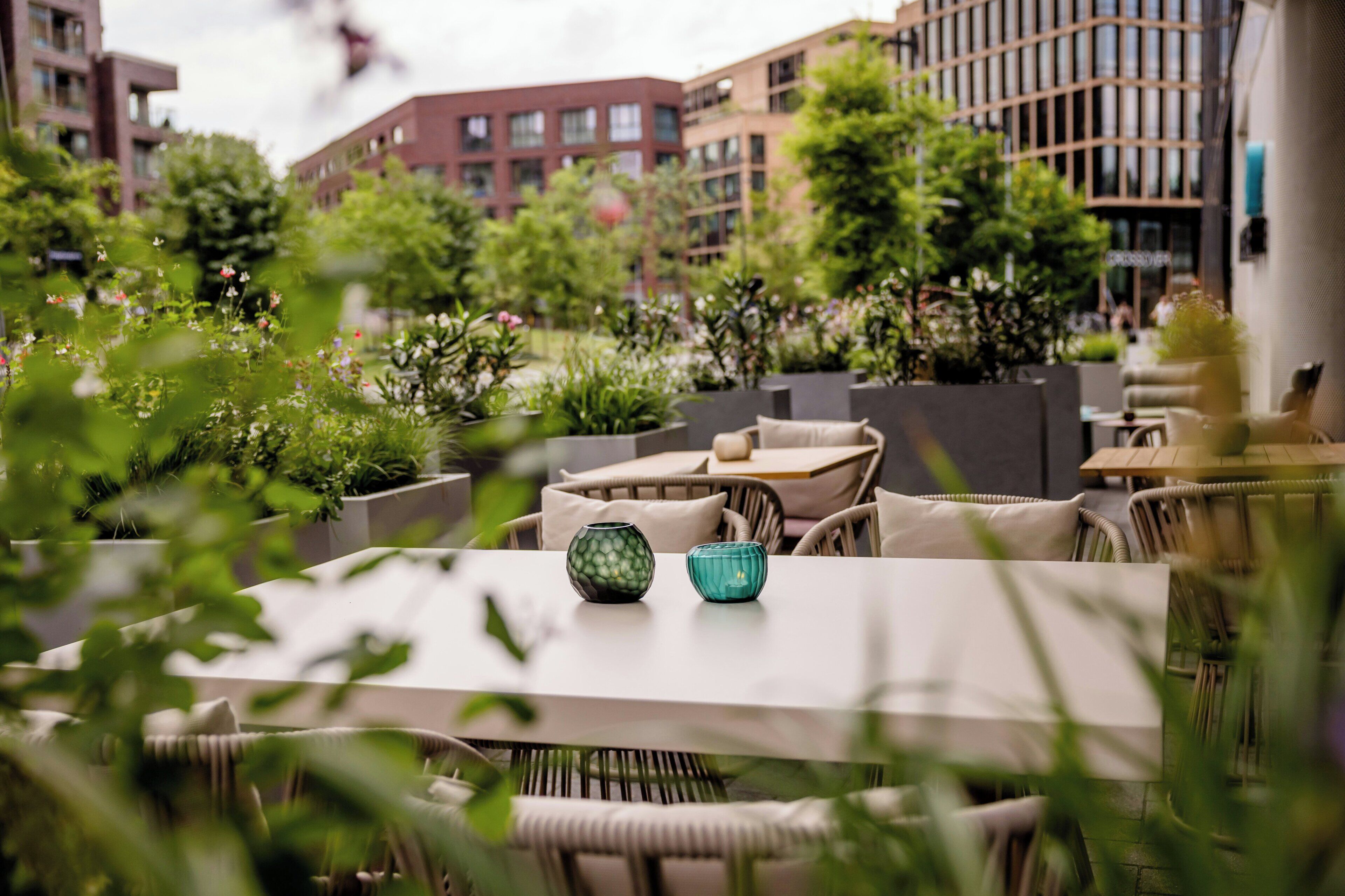 An outdoor seating area with tables and chairs, surrounded by greenery and buildings in the background.