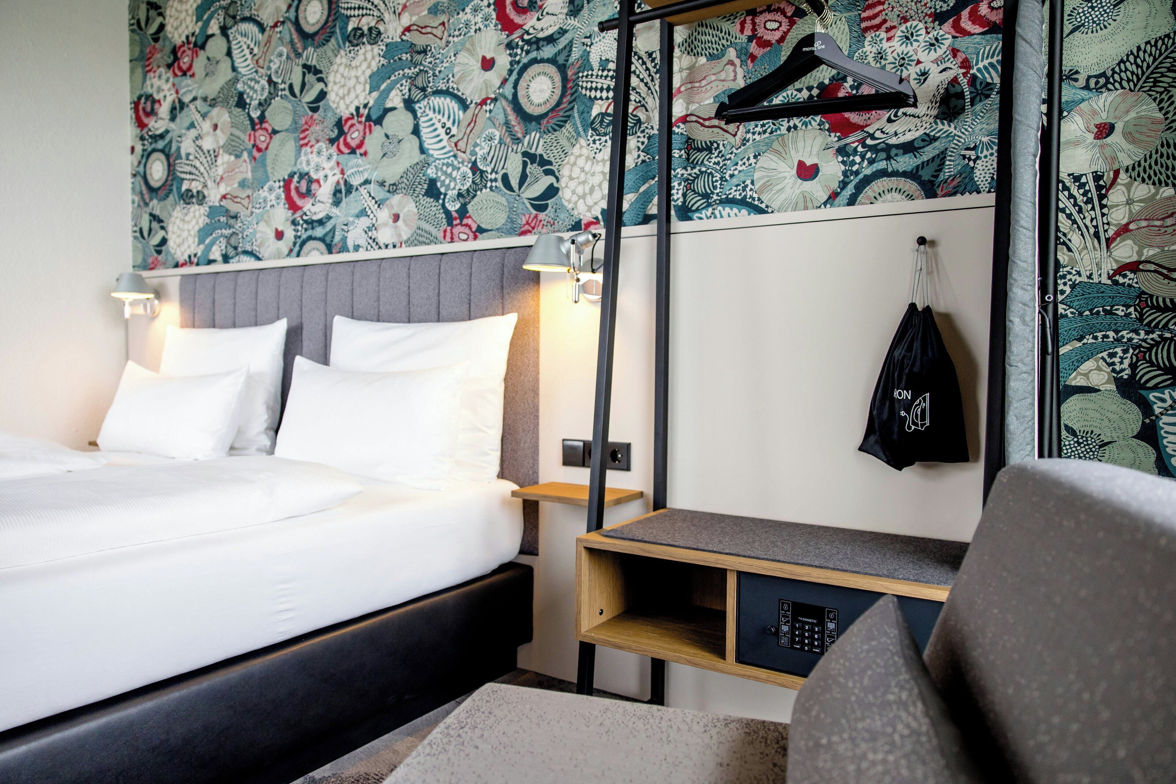 A hotel room with a bed, bedside table, lamp, and a wall with a patterned wallpaper.