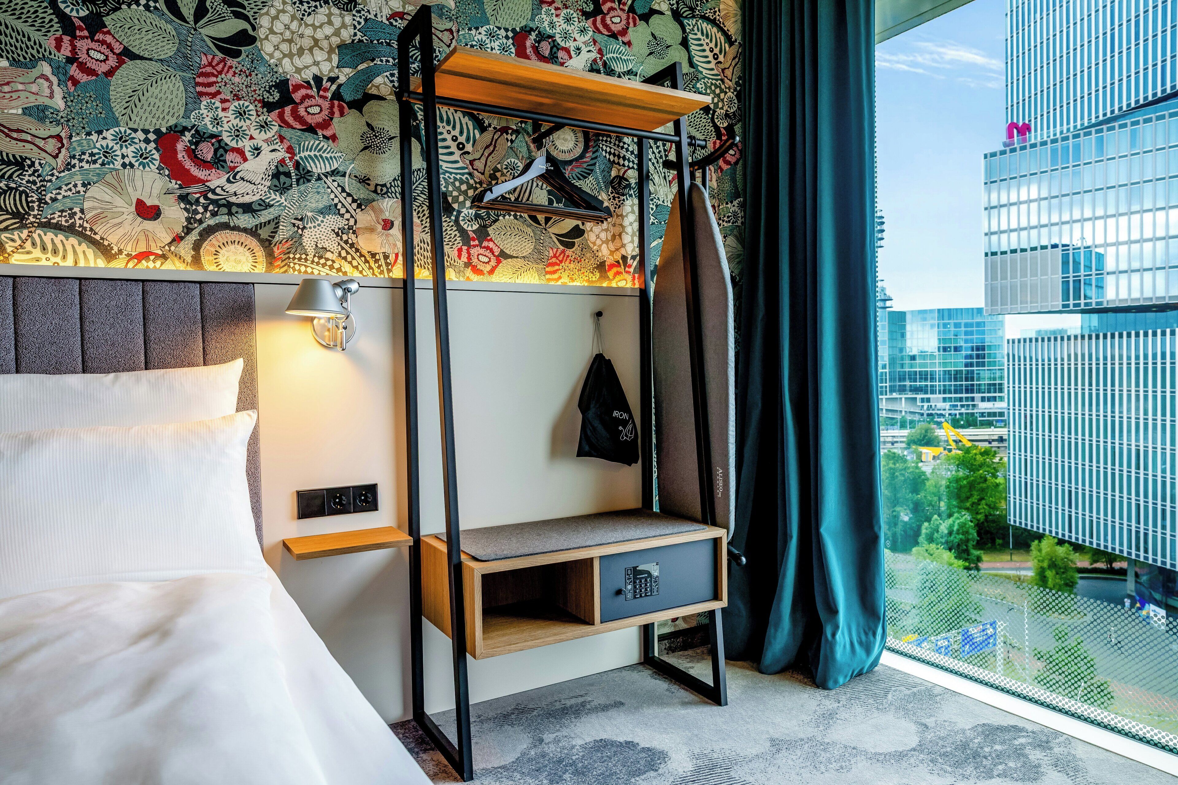 Hotelkamer met een bed, een bedlampje, een plank met een tas en uitzicht op een stadslandschap door een raam.