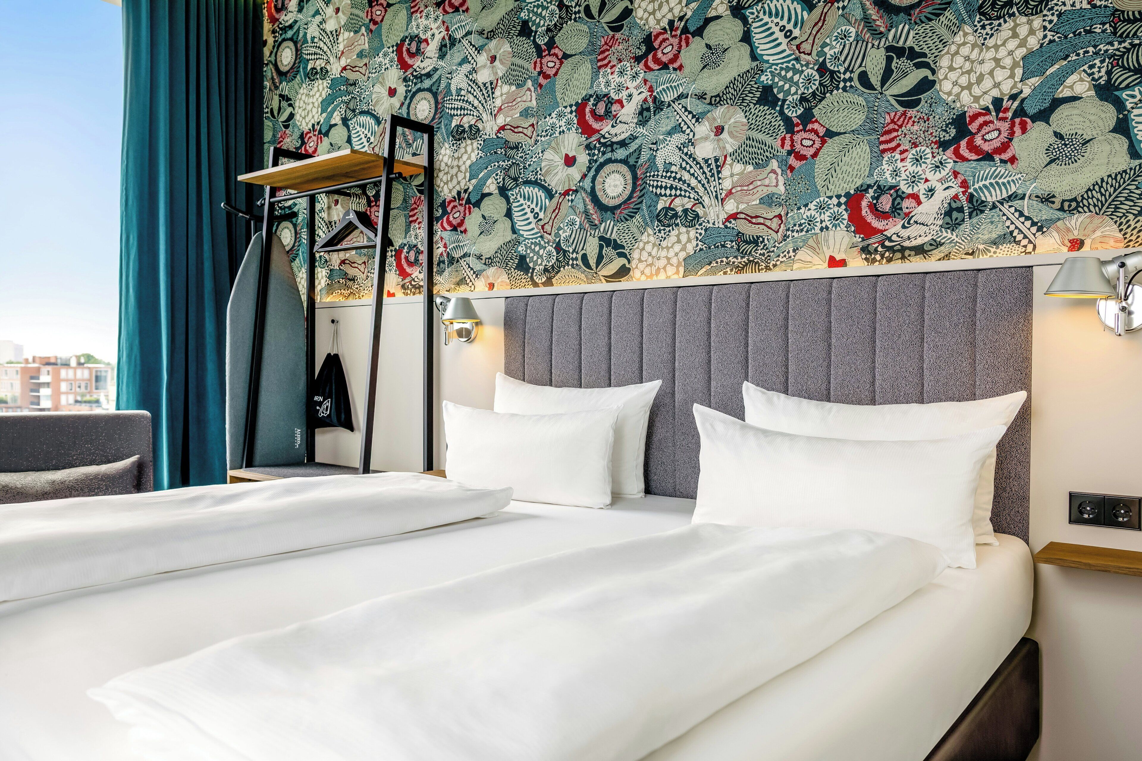 Quarto de hotel com uma cama grande, cabeceira com padrão decorativo, mesa de cabeceira e janela com cortinas.