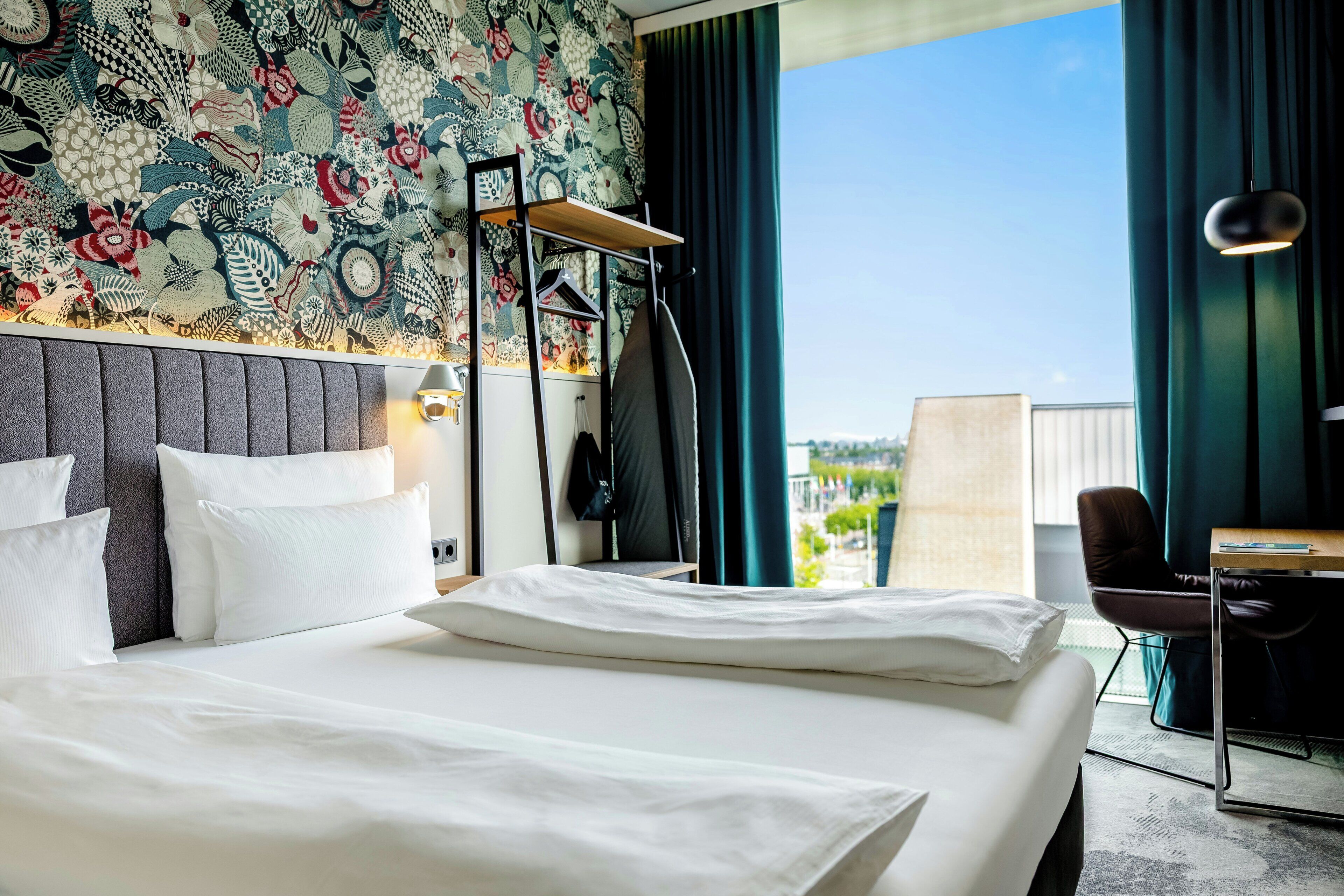 A hotel room with a large bed, a desk, a chair, and a view of the city.