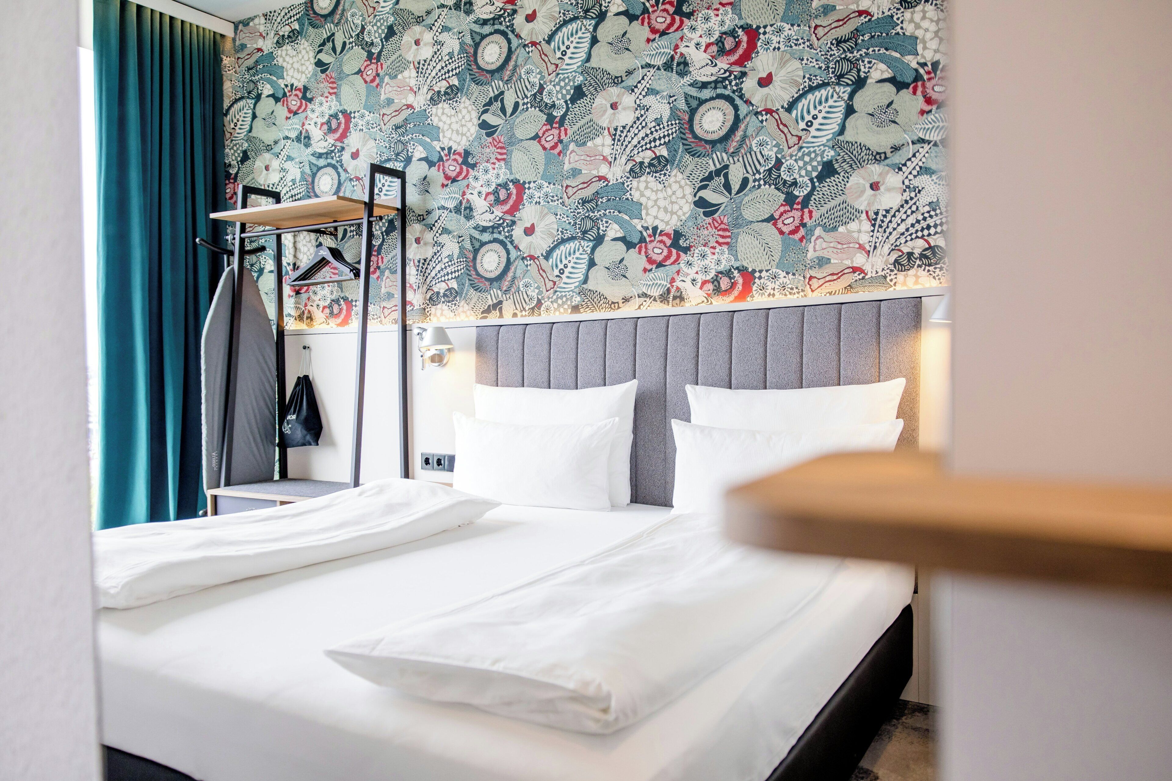A hotel room with a neatly made bed, a hanging coat rack, and a patterned wallpaper.