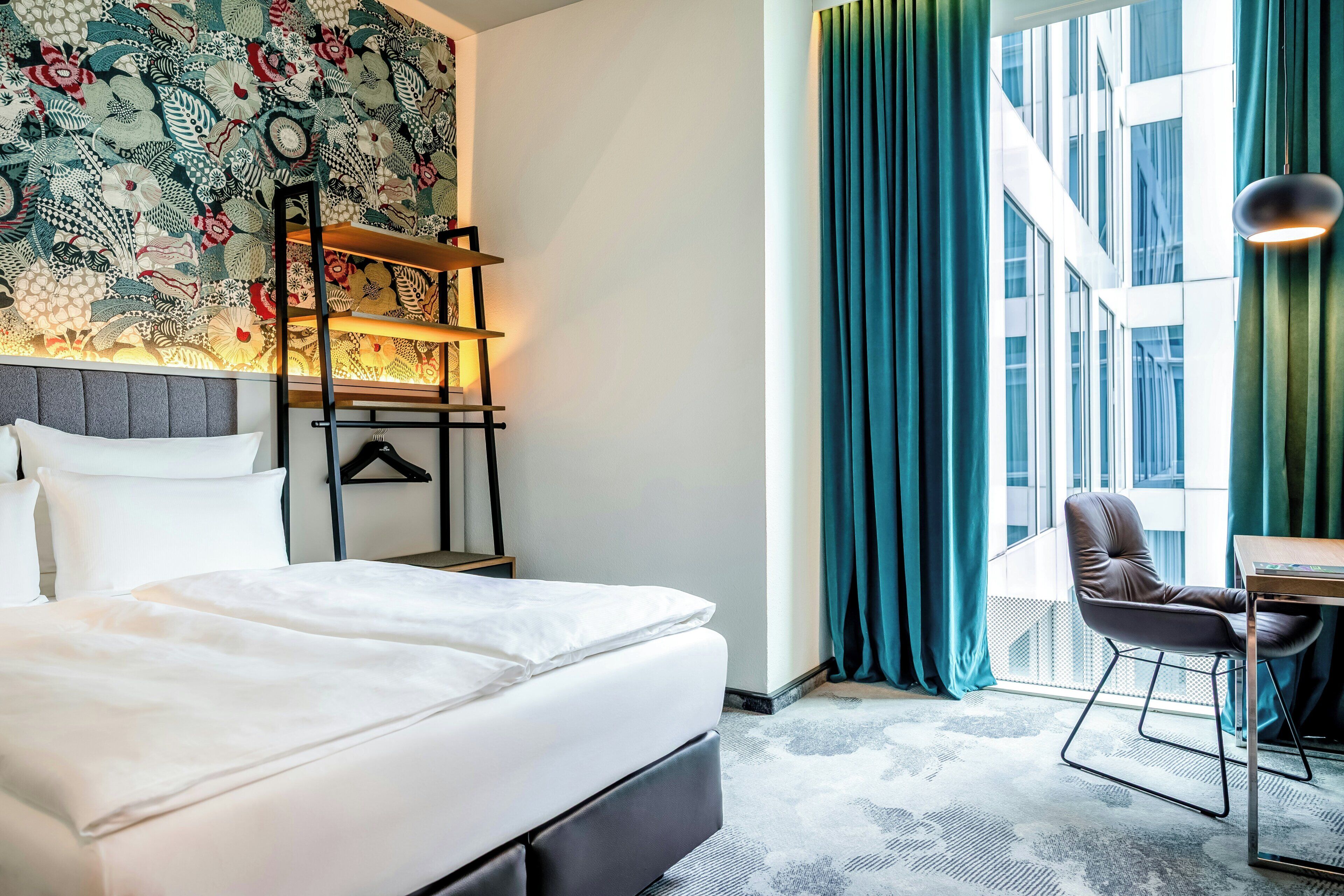 A hotel room with a bed, a desk, a chair, a window with curtains, and a wall with a patterned tapestry.
