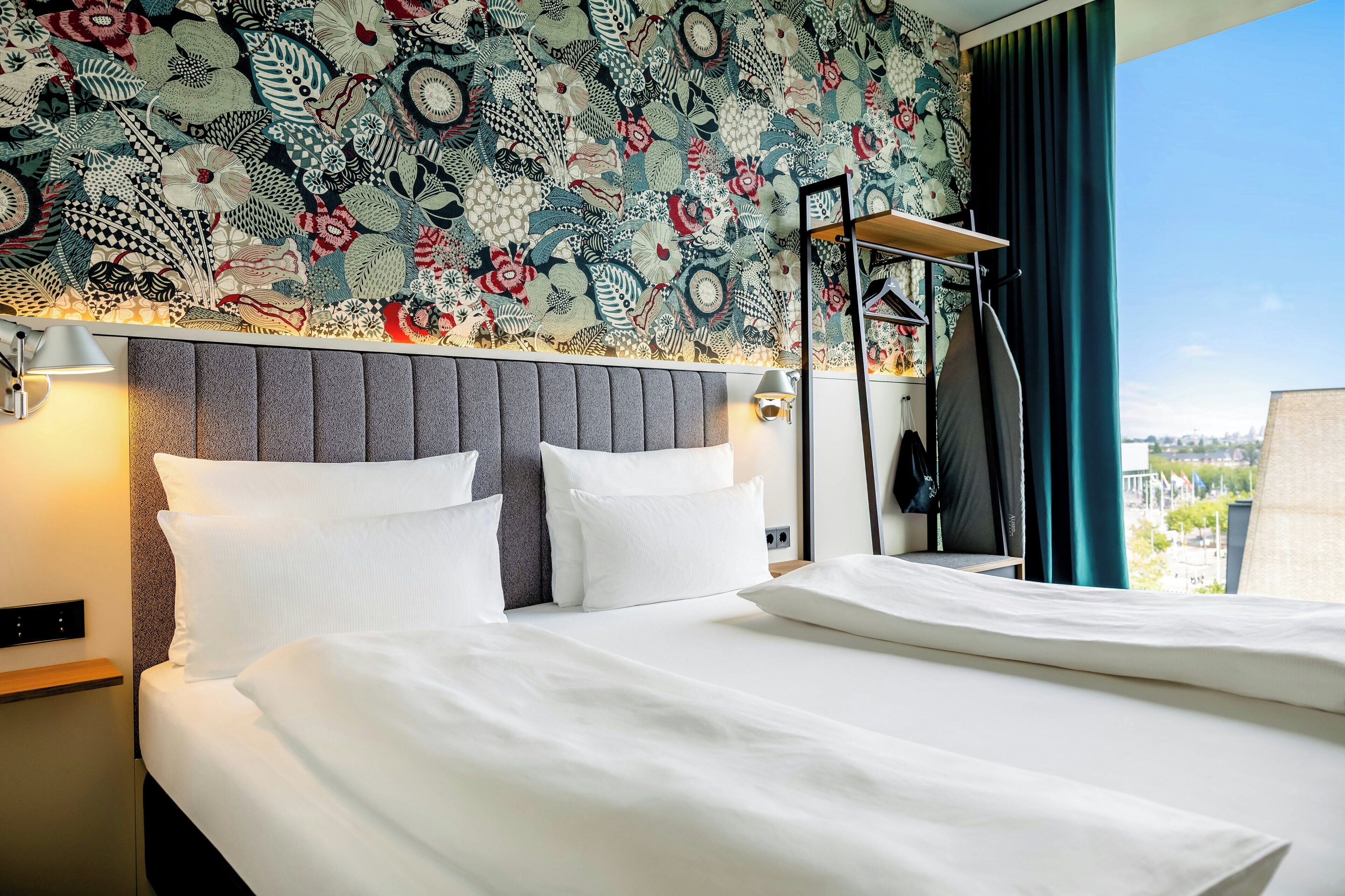 A hotel room with a bed, pillows, a headboard, a nightstand, a lamp, a coat rack, and a window with a view.
