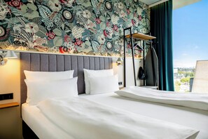 A hotel room with a bed, pillows, a headboard, a nightstand, a lamp, a coat rack, and a window with a view.