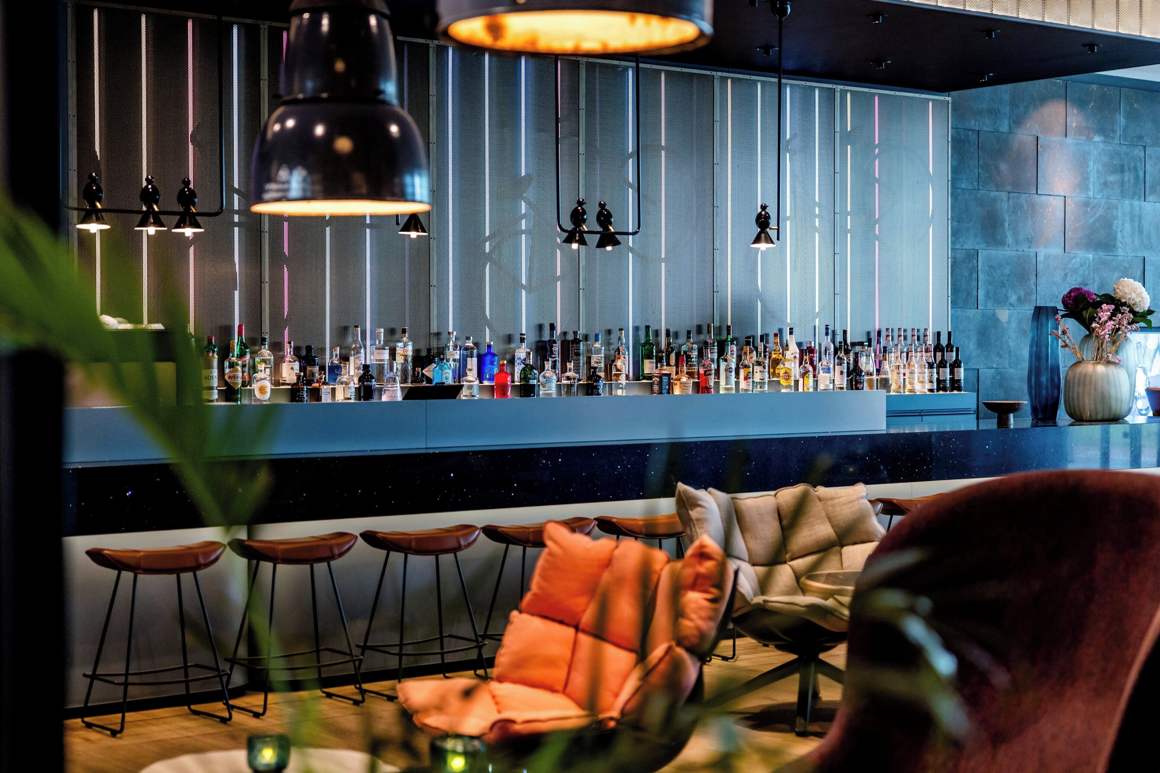 Uma área de bar com diversos tipos de garrafas, banquetas e iluminação pendente.