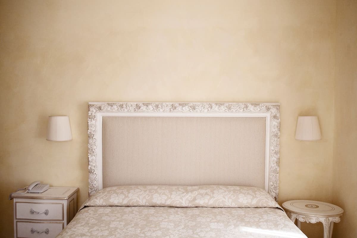 Un lit avec une tête de lit blanche et une literie à motifs floraux, une table de chevet avec une lampe et une petite table de nuit avec un téléphone.