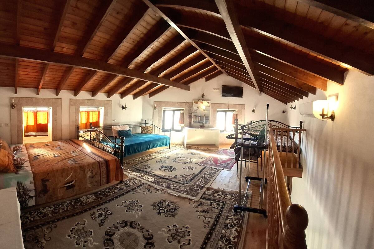A spacious attic room with a high wooden ceiling, a large bed with a blue bedspread, a staircase, and a view of the countryside.