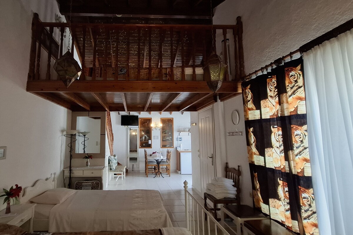 A bedroom with a bed, a small table, and a chair. There is a balcony with a wooden railing and a view of the outside.
