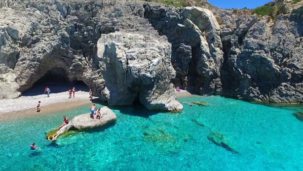 A rocky coastal area with clear turquoise water, a cave entrance, and people engaging in water activities. - Castellino Studios (Rhodes)