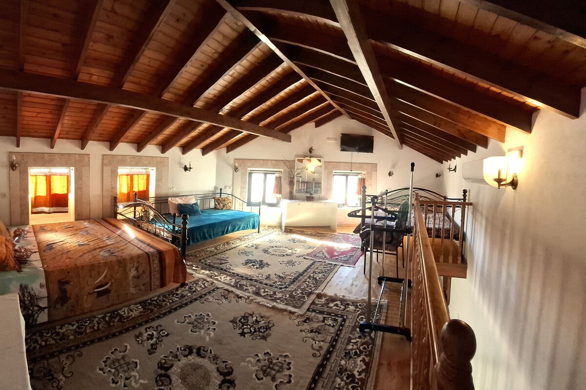 A spacious attic room with a high wooden ceiling, a large bed with a blue bedspread, a staircase, and a view of the countryside.