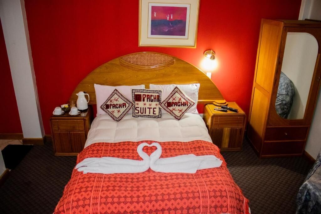 A hotel room with a red wall, a wooden headboard, a bed with a red patterned bedspread, and two pillows with 'Pachacuti' branding.