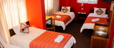 A hotel room with three single beds, each with a red patterned bedspread and white pillows. There are wooden headboards and a small wooden table between the beds. A television is mounted on the wall above the beds.