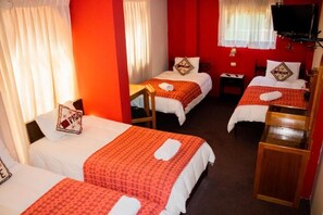 A hotel room with three single beds, each with a red patterned bedspread and white pillows. There are wooden headboards and a small wooden table between the beds. A television is mounted on the wall above the beds. - Pacha Suite (Puno)