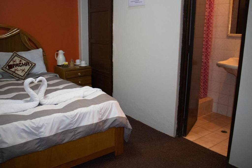 Ett hotellrum med en säng, ett nattduksbord, en träpanel vid sänggaveln, en konstnärligt arrangerad handduk på sängen, ett litet bord med en kanna och glas, och ett badrum som syns genom en öppen dörr.