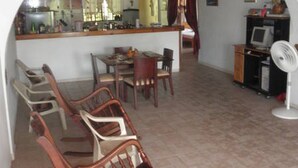 A dining area with wooden chairs and a table, a kitchen with a counter and appliances, and a fan on the floor. - Ground Road Native Place (San Andrés)