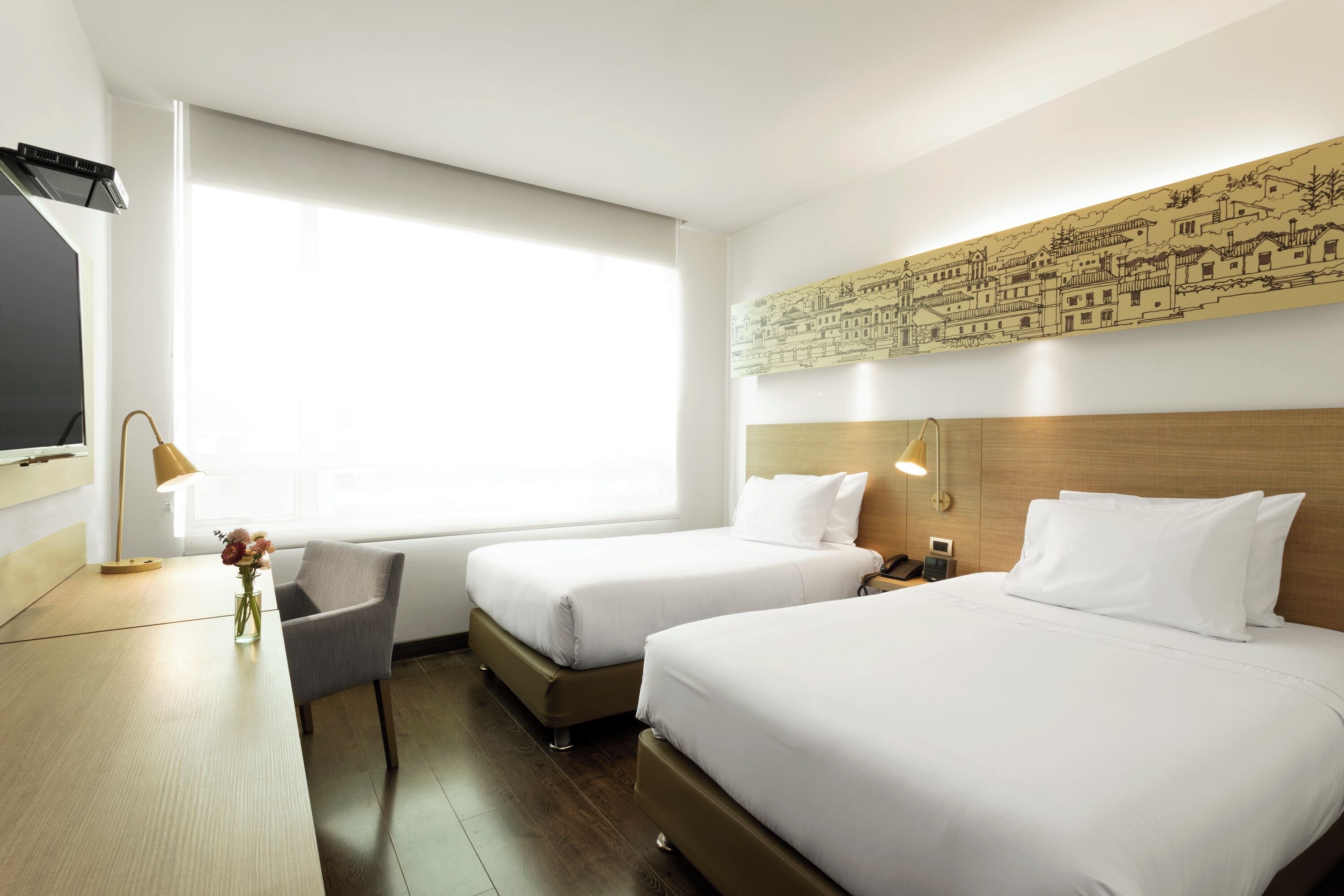 A hotel room with two beds, a desk, a lamp, a vase with flowers, and a large window.