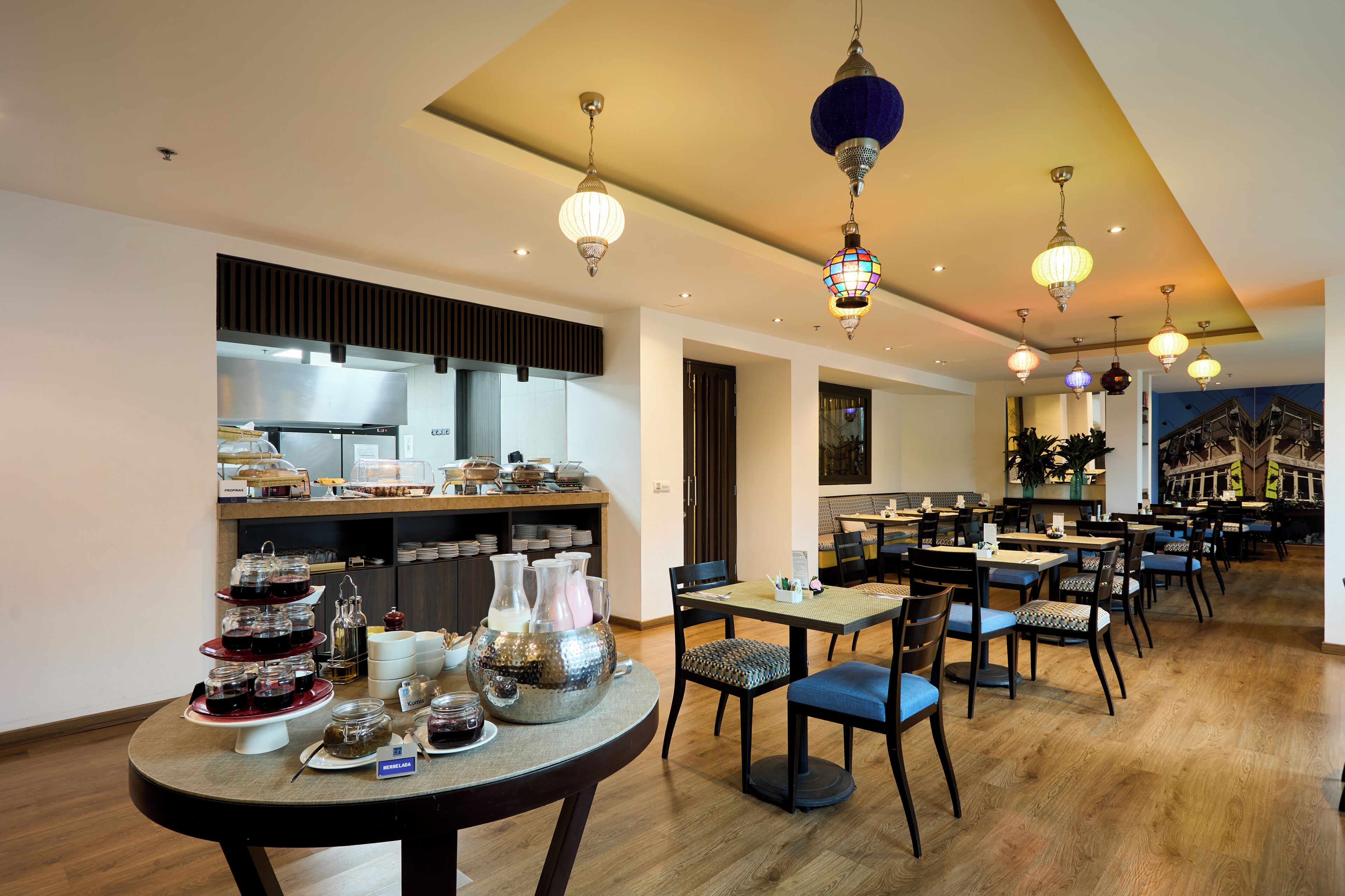 A modern dining area with multiple tables and chairs, a kitchen in the background, and decorative hanging lights.