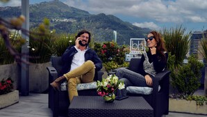 A couple seated on wicker furniture with a view of a mountain and a building in the background.