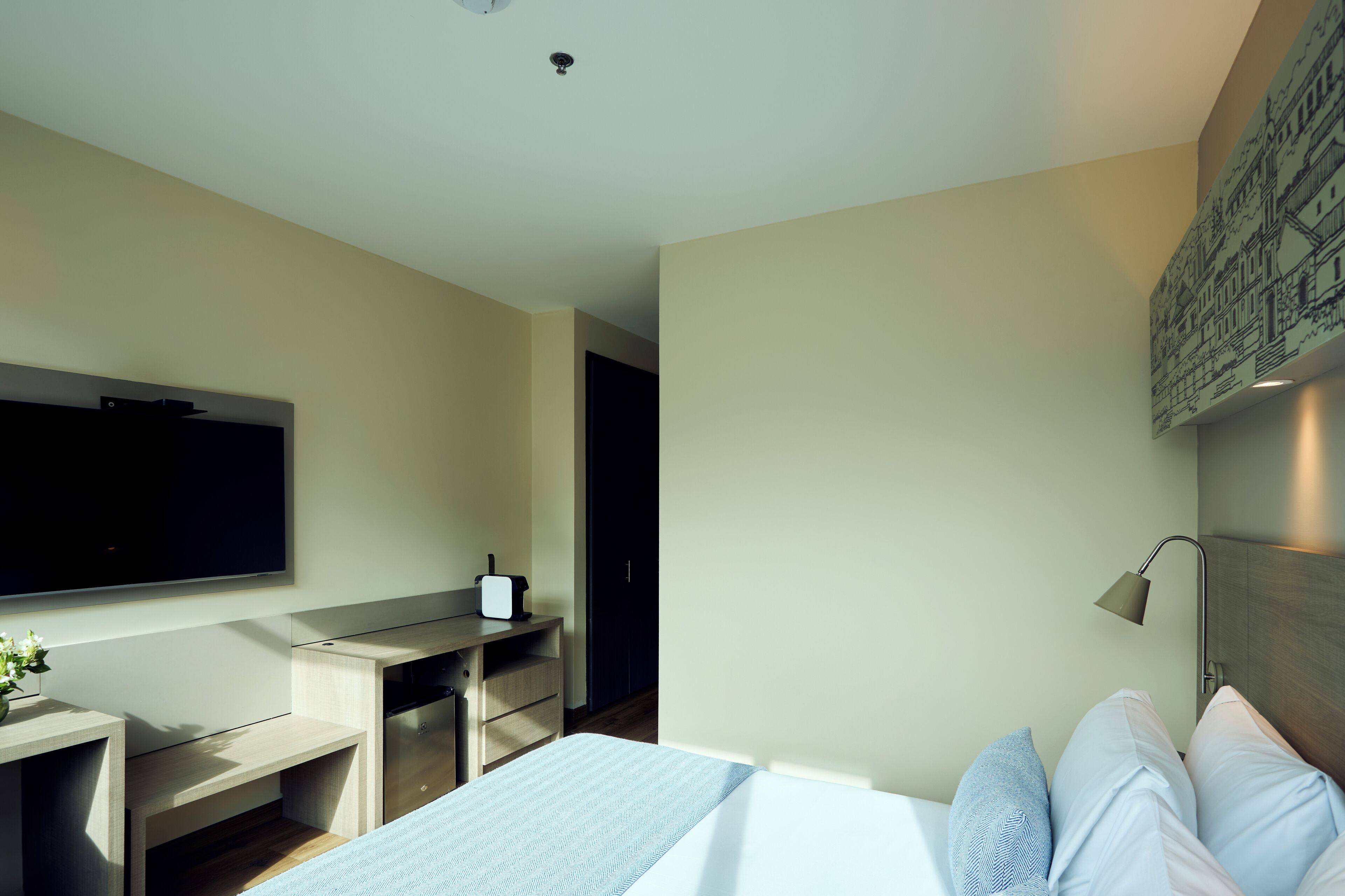 A modern hotel room with a large bed, a desk with a flat-screen TV, a small table with a vase, and a wall-mounted lamp.