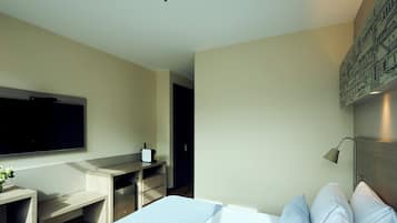 A modern hotel room with a large bed, a desk with a flat-screen TV, a small table with a vase, and a wall-mounted lamp.