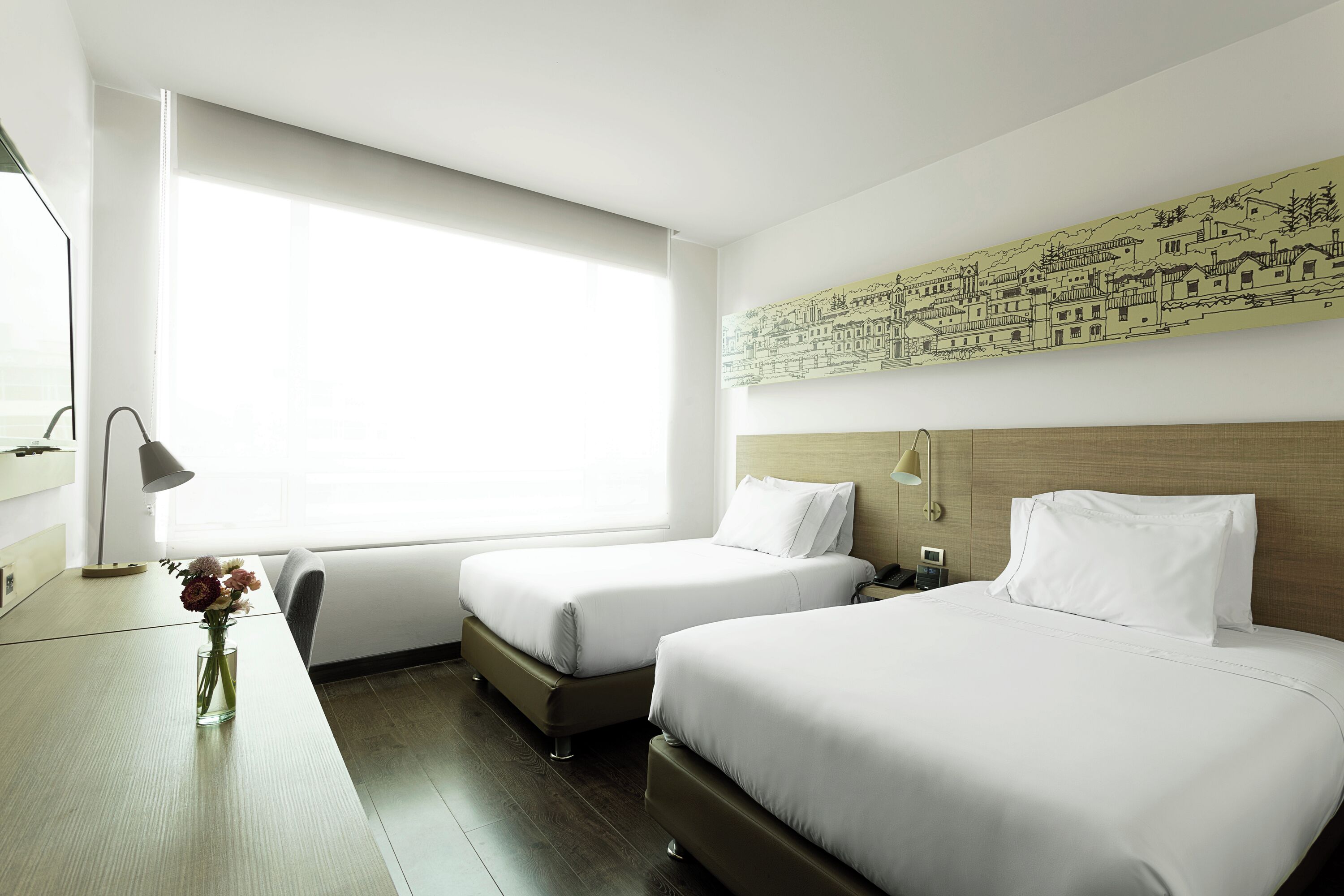 Habitación de hotel con dos camas, un escritorio, un florero con flores y un ventanal grande.