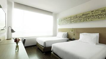 Habitación de hotel con dos camas, un escritorio, un florero con flores y un ventanal grande.