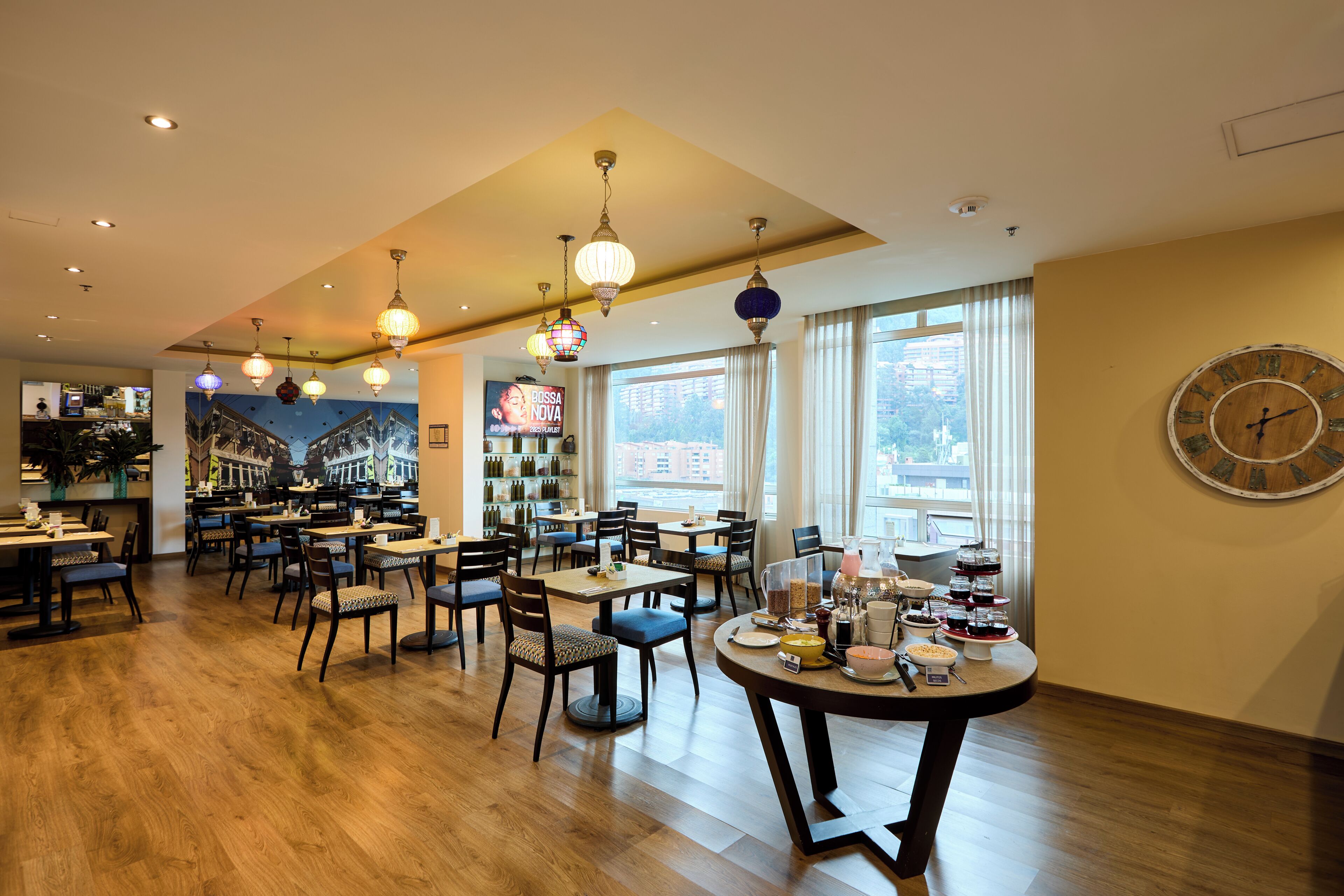 Een restaurant met meerdere eettafels en stoelen, een klok aan de muur en uitzicht op de stad door grote ramen.