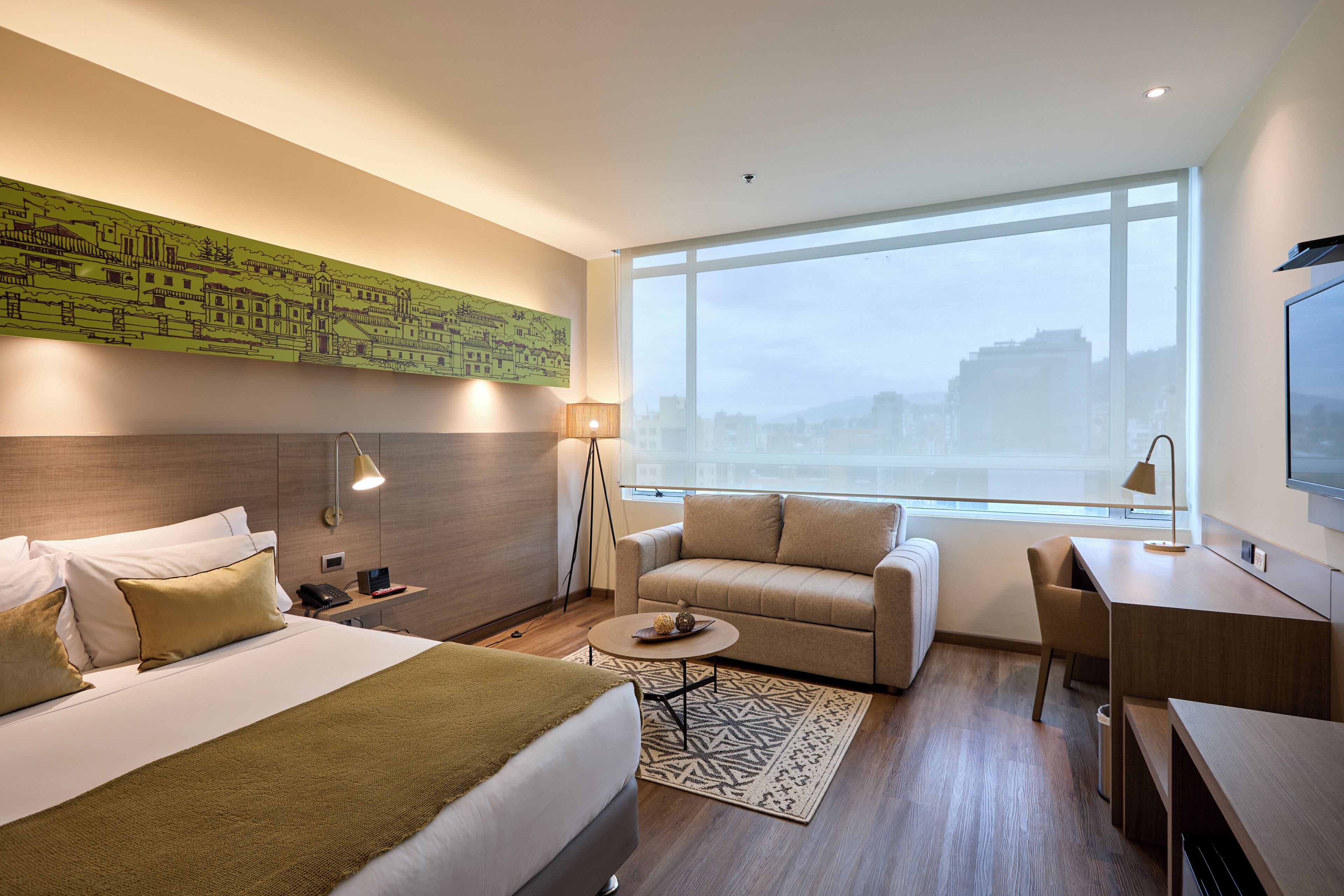 A hotel room with a bed, a sofa, a desk, a chair, a small table, a lamp, a TV, and a large window with a view.