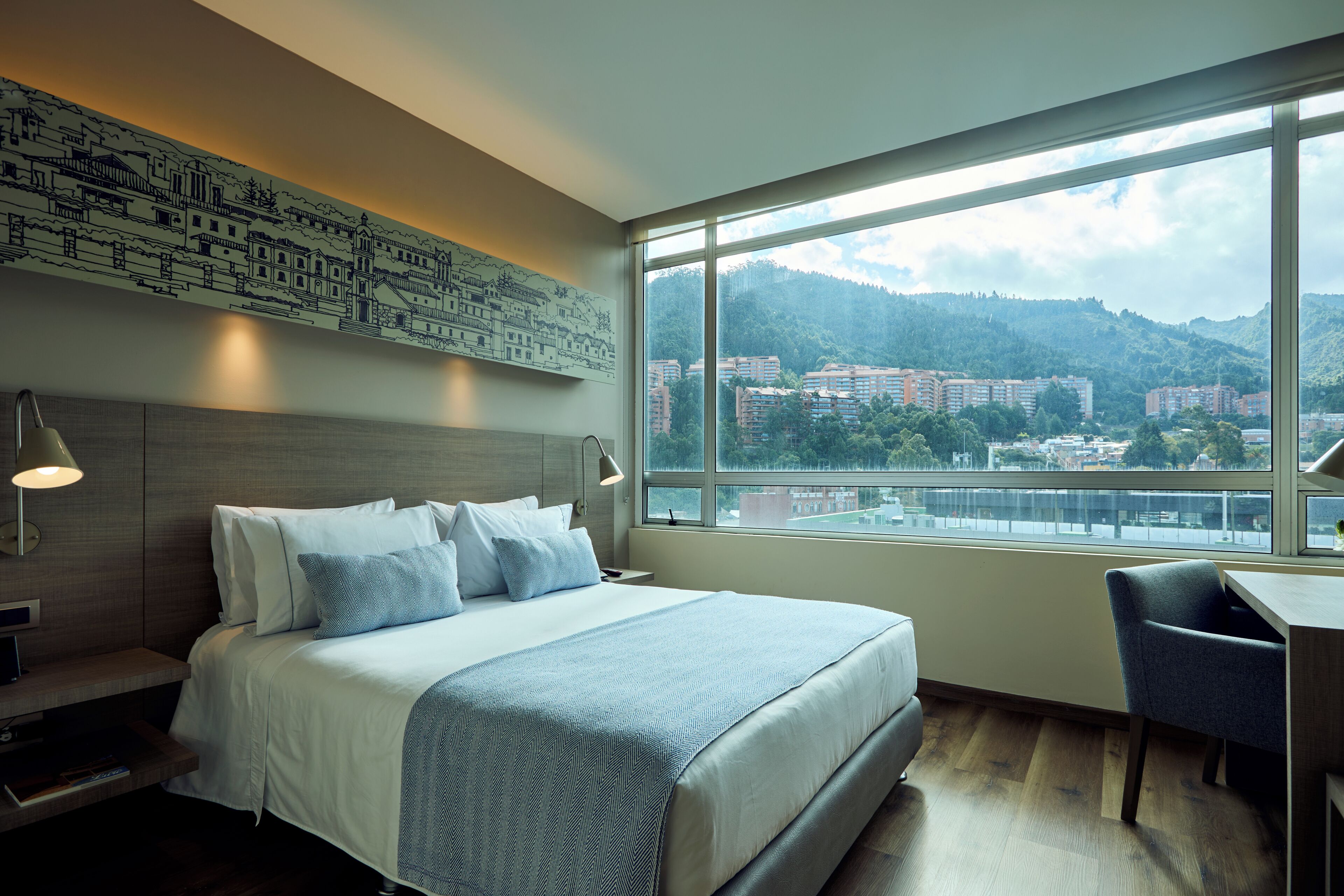 Habitación de hotel con una cama grande, un escritorio y vistas a un paisaje urbano a través de un amplio ventanal.
