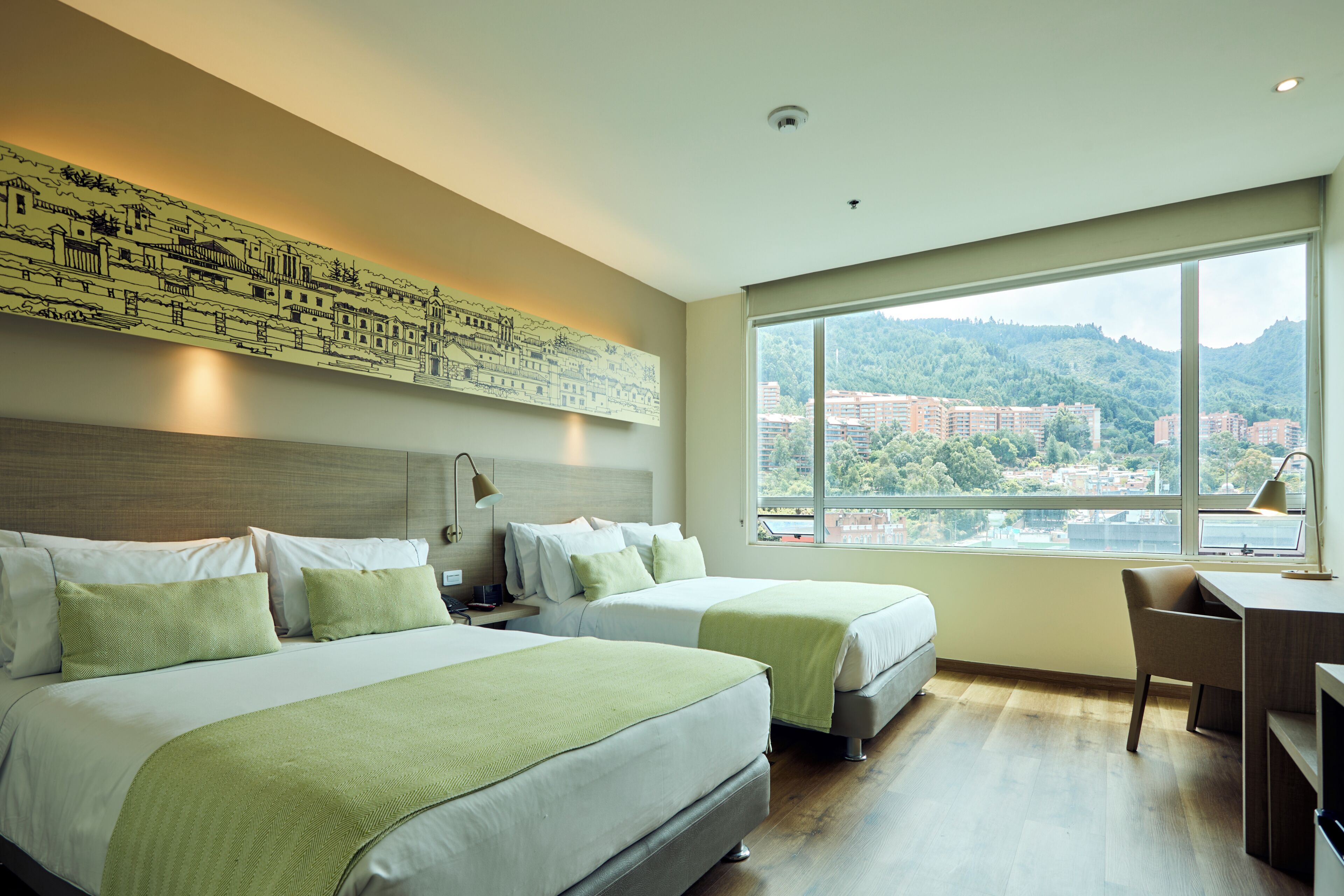 Ein Hotelzimmer mit zwei Betten, einem Schreibtisch und einem großen Fenster mit Blick auf die Stadt.